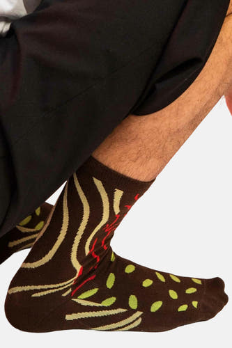 Chaussettes brunes d'EAT MY SOCKS avec des lignes ondulées, des taches et des détails rouges, portées avec un pantalon noir.