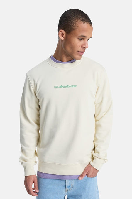Ecru sweater met ronde hals van Quiksilver, met groene merknaam geborduurd op de borst, gedragen over een lila shirt, met lichte jeans.