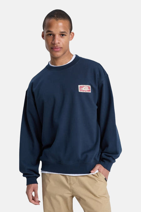 Blauwe sweater met ronde hals van Quiksilver, met een rechthoekige patch op de borst, gedragen met een wit shirt en khaki broek.

