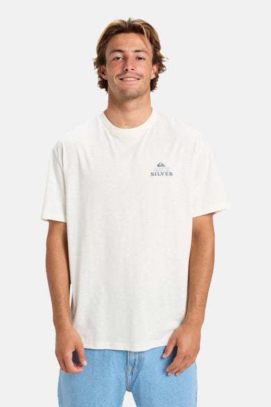 T-shirt écru à manches courtes de Quiksilver, avec un petit logo sur la poitrine et porté avec un pantalon de travail bleu.