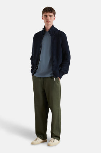 T-shirt bleu à manches courtes de FARAH, porté sous un gilet bleu marine avec fermeture éclair, associé à des pantalons verts et des chaussures crème.