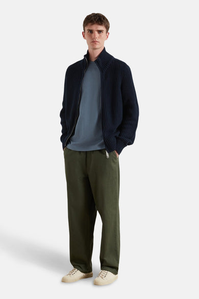 T-shirt bleu à manches courtes de FARAH, porté sous un gilet bleu marine avec fermeture éclair, associé à des pantalons verts et des chaussures crème.