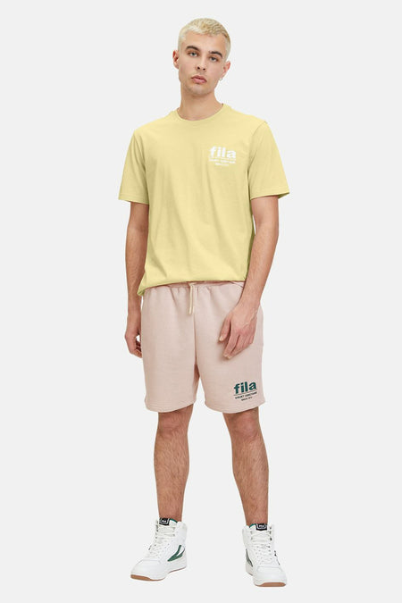 T-shirt jaune à manches courtes de FILA, avec une impression blanche sur la poitrine, associé à des shortses roses et des baskets blanches.