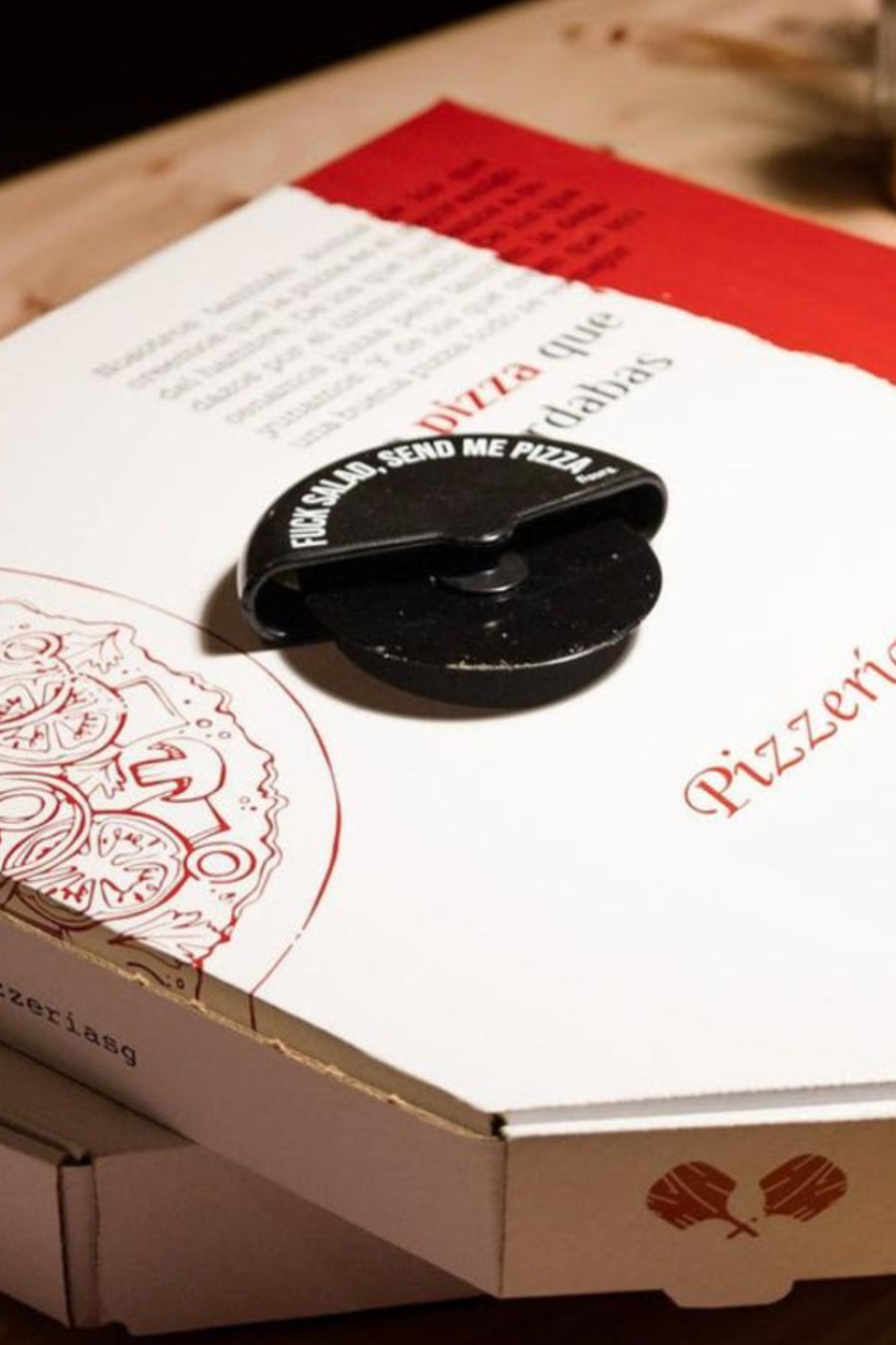 Zwarte pizzasnijder met de tekst "FUCK SALAD, SEND ME PIZZA" op een witte pizzadoos.