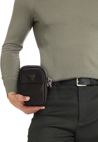 Sac à dos noir de Guess, avec un logo argenté et une fermeture éclair, porté avec un pull olive et un pantalon rayé.