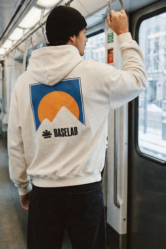 Witte sweater met kap van BASELAB, met een blauw en oranje logo op de rug, gedragen met zwarte jeans.
