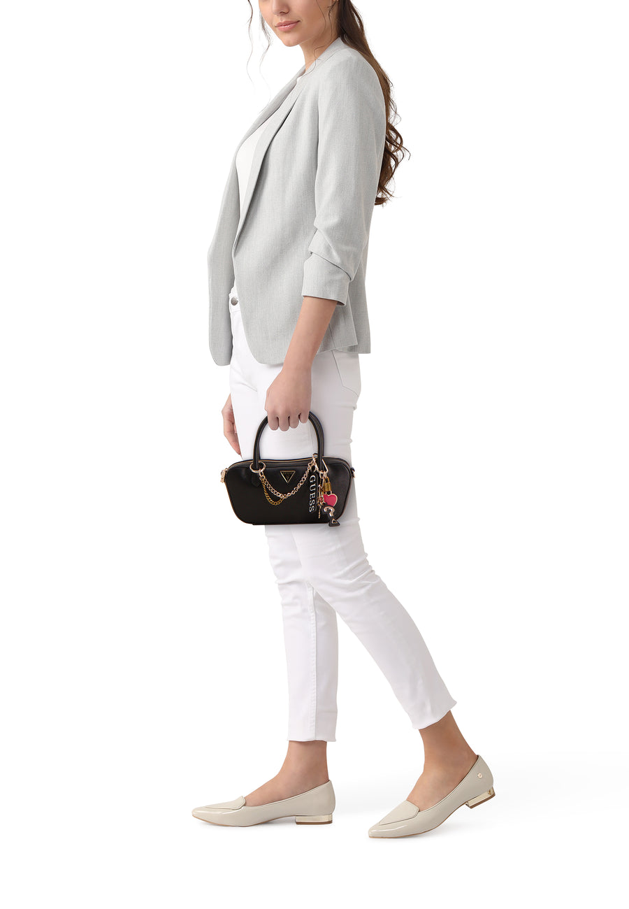 Sac à main noir Guess, avec détails dorés et breloques, porté avec un blazer gris et un pantalon blanc.