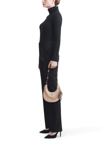 Sac à bandoulière beige Guess avec détails de chaîne dorée et breloque de marque noire, porté par une femme en col roulé noir et pantalon.