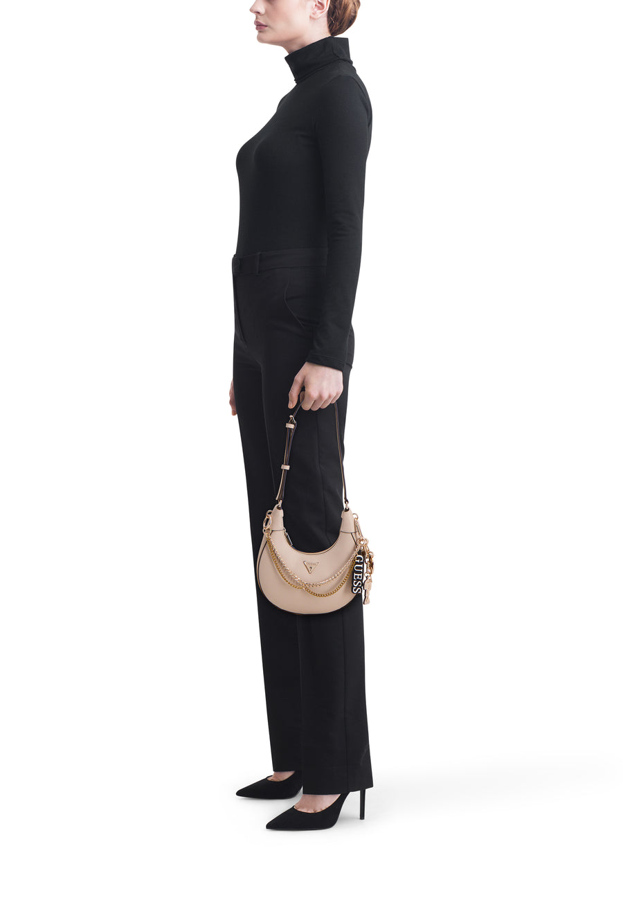 Sac à bandoulière beige Guess avec détails de chaîne dorée et breloque de marque noire, porté par une femme en col roulé noir et pantalon.