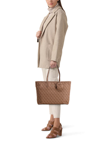 Sac à dos marron de Guess avec un motif monogramme, porté par un modèle en une veste beige et des sandales marrons.