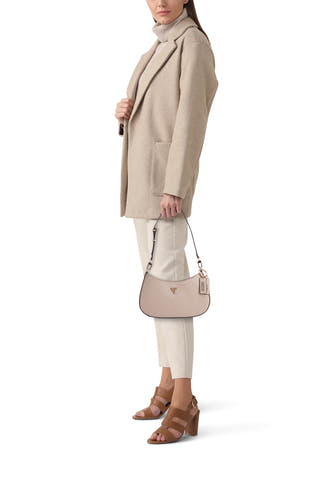 Sac à bandoulière beige de Guess, avec pendentif logo, porté avec un manteau beige, un pantalon blanc et des sandales marron.