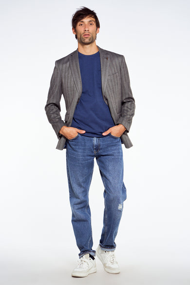 Homme en blazer gris, chemise bleue, jeans et baskets blanches, les mains dans les poches.