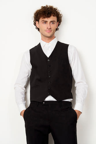 Zwart gilet van Le Fabuleux Marcel de Bruxelles, gedragen over een wit overhemd en zwarte broek, met handen in de zakken.