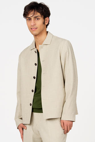 Blazer beige de Le Fabuleux Marcel de Bruxelles, avec fermeture à boutons et fines rayures verticales, porté sur un pull vert.