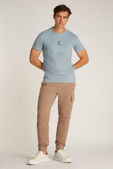 T-shirt bleu clair à manches courtes de Calvin Klein Jeans, porté avec un pantalon de jogging marron à poches et des baskets blanches.