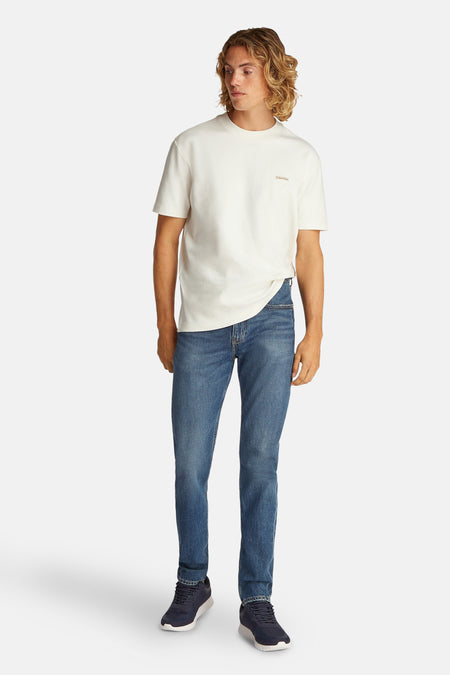 T-shirt écru à manches courtes de Calvin Klein, porté avec des jeans bleus et des baskets bleu marine.
