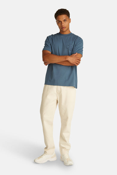 T-shirt bleu à manches courtes de Calvin Klein, avec des bras croisés et un pantalon beige et des baskets.