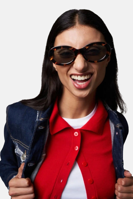 Lunettes de soleil marron de KOMONO, avec une monture écaille de tortue, portées avec un chemisier rouge et une veste en jean.