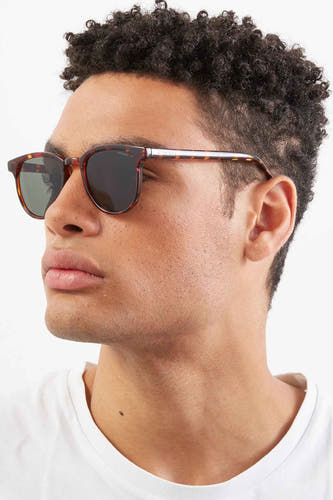 Lunettes de soleil vertes de KOMONO, portrait rapproché d'un homme aux cheveux bouclés et à un T-shirt blanc.