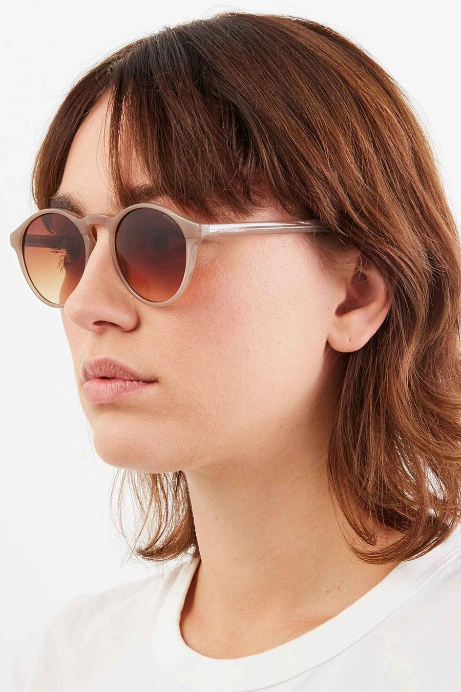 Lunettes de soleil beige de KOMONO, avec une lentille graduée marron-jaune et portées par une femme en un chemisier blanc.