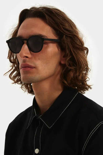 Lunettes de soleil noires de KOMONO, portées par une personne avec un chemisier noir à des boutons blancs.