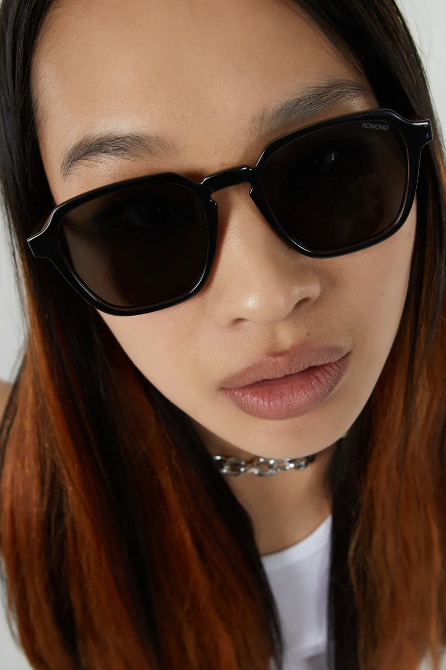 Lunettes de soleil noires de KOMONO, portées avec une chaîne de couleurs argentées et un t-shirt blanc.