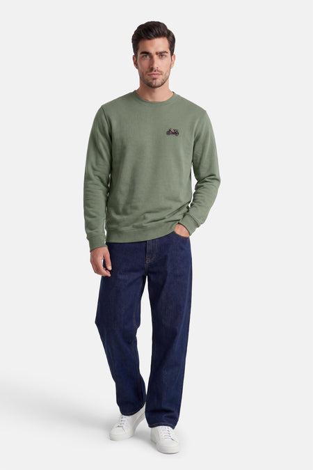 Pull vert à col rond de LAKOR, avec une petite moto brodée sur la poitrine, porté avec un jean bleu foncé et des baskets blanches.