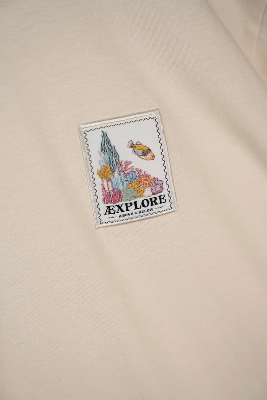 T-shirt écru à manches courtes de LAKOR, avec un patch carré à motif sous-marin et le texte "Explore".