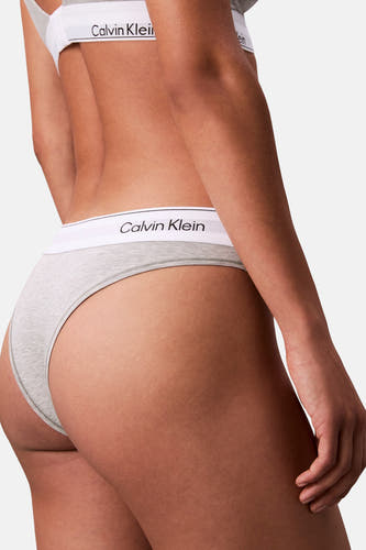 Grijze string van Calvin Klein, close-up achterkant met witte logoband en zichtbare hand.
