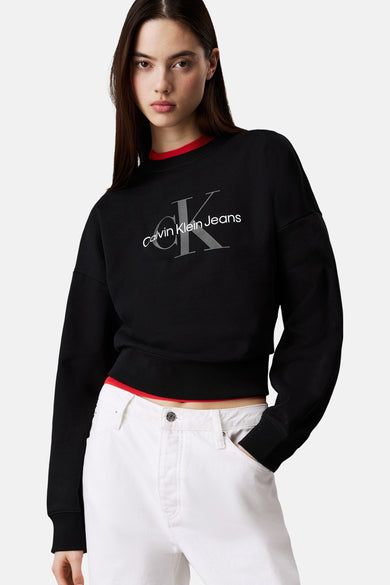Pull-over noir avec col rond de Calvin Klein Jeans, avec le nom de la marque gris sur l'avant, porté avec un pantalon blanc.