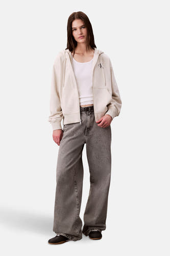 Gilet beige de Calvin Klein, avec des manches longues et une fermeture éclair, associé à un chemisier blanc et des jeans gris larges.
