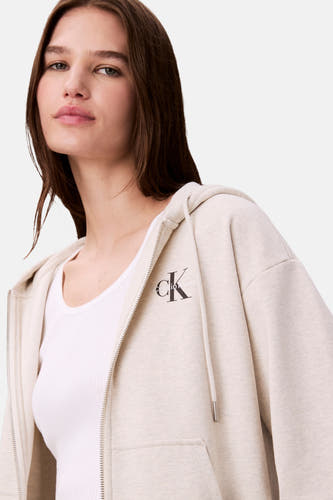 Gilet beige de Calvin Klein avec capuchon et fermeture à glissière à l'avant, porté sur une chemise blanche.
