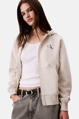 Veste beige de Calvin Klein, avec fermeture éclair et capuche, portée sur un chemisier blanc avec une ceinture noire.