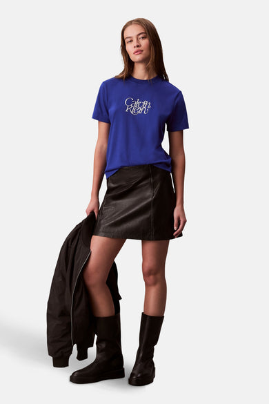 T-shirt bleu à manches courtes de Calvin Klein, porté avec une jupe en cuir noire et des bottes, tenant un manteau noire dans la main.