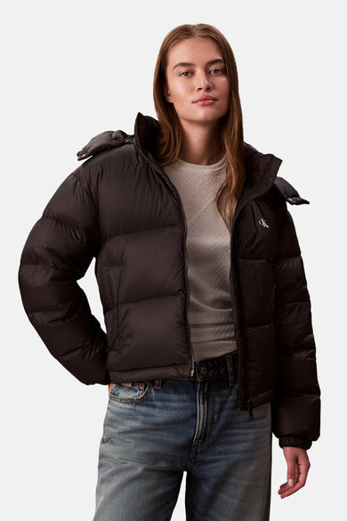 Manteau court noir de Calvin Klein, avec une texture remplie de duvet et un logo sur la poitrine, porté avec un pull beige et des jeans.