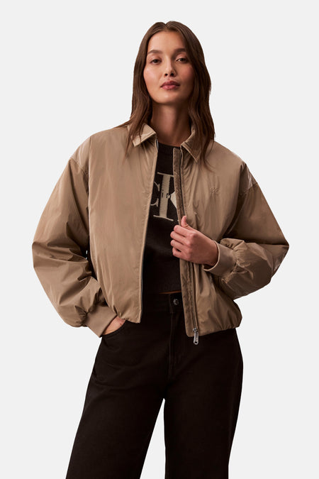 Manteau court beige de Calvin Klein, avec fermeture éclair visible et porté sur une chemise et un pantalon noirs.