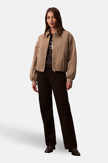 Manteau court beige de Calvin Klein, avec des manches longues, porté avec un pull sombre et un pantalon noir.