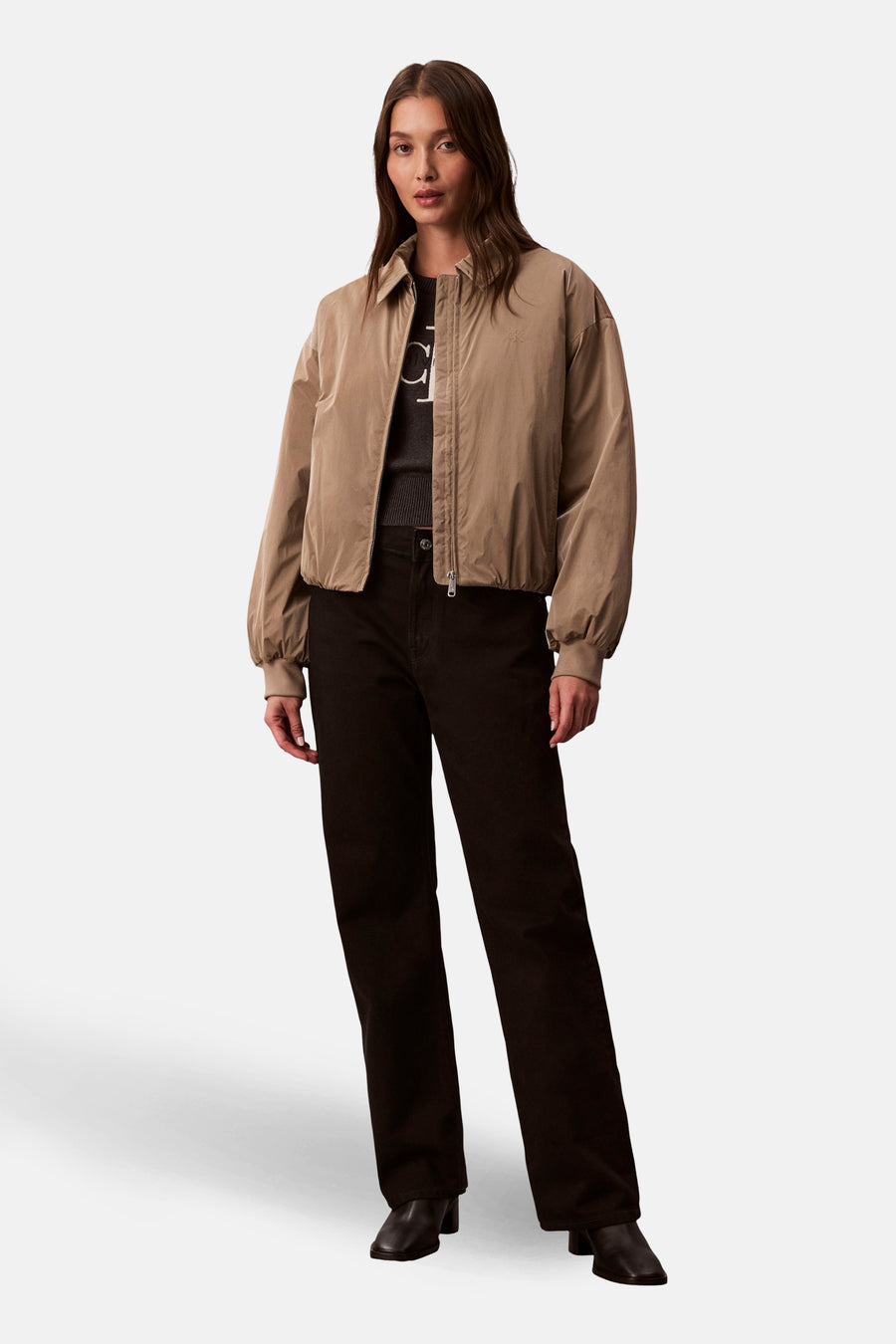 Manteau court beige de Calvin Klein, avec des manches longues, porté avec un pull sombre et un pantalon noir.