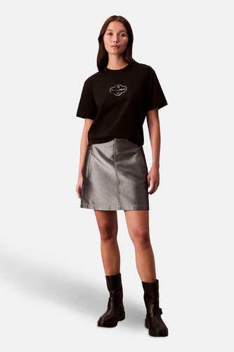 T-shirt noir à manches courtes de Calvin Klein, avec logo sur la poitrine, porté avec une jupe argentée et des bottes noires.