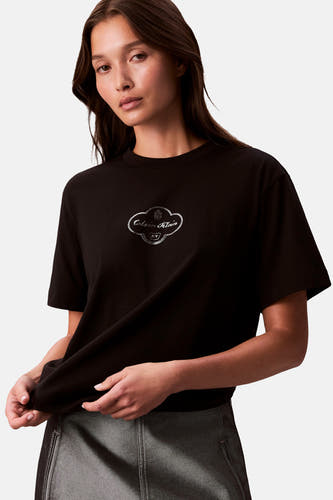 T-shirt noir à manches courtes de Calvin Klein, avec logo sur la poitrine et porté avec un pantalon gris.