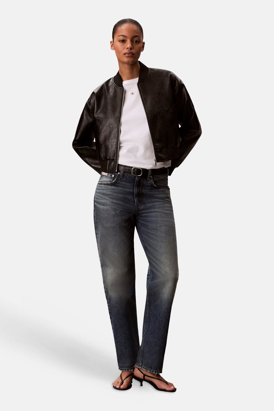 T-shirt blanc Calvin Klein, à manches courtes, porté sous un blouson bomber en cuir noir, associé à un jean bleu et des sandales noires.