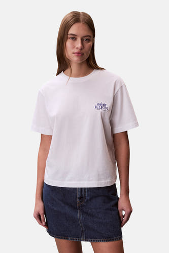 T-shirt blanc à manches courtes Calvin Klein, avec un petit logo violet sur la poitrine et porté avec une jupe en jean bleue.