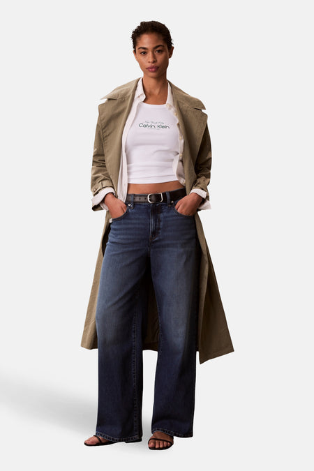 Débardeur blanc Calvin Klein, avec col rond et imprimé logo, combiné avec un trench kaki, une chemise blanche et un jean foncé.