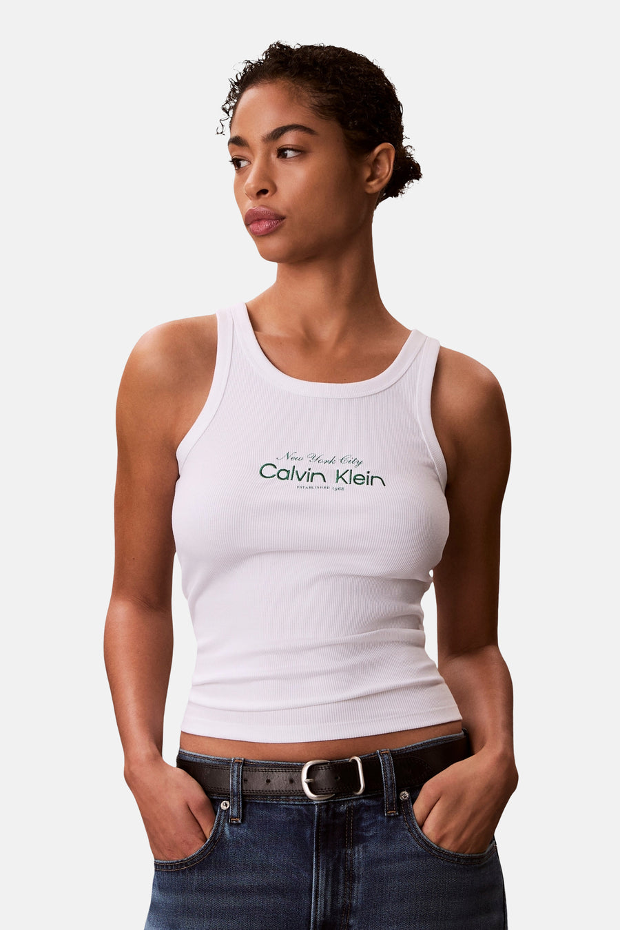 Débardeur blanc Calvin Klein, avec texture côtelée et un logo vert foncé sur la poitrine, porté avec un jean et une ceinture.