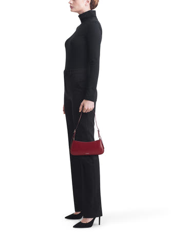 Femme avec un pull noir et une jupe, une poche rouge, des escarpins noirs, debout.