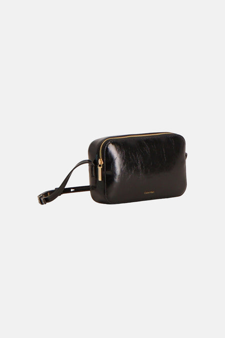 Sac à bandoulière noir brillant Calvin Klein avec fermeture éclair dorée, bandoulière réglable et petit logo, présenté en biais.