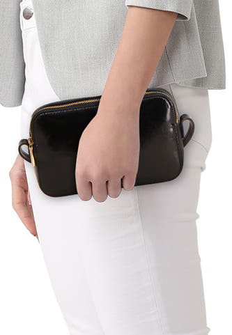 Sac à bandoulière noir brillant de Calvin Klein avec fermeture éclair dorée et petites poignées, contre la hanche d'une personne en blazer gris et pantalon blanc.