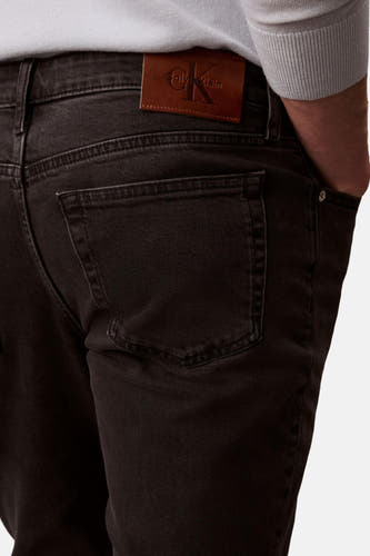 Jean slim fit noir de Calvin Klein, avec une étiquette en cuir marron, un poche arrière et une main dans la poche.