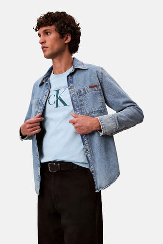 T-shirt bleu à manches courtes de Calvin Klein, porté sous un pull en denim ouvert et avec un pantalon noir.