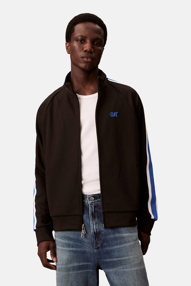 Pull-over noir de Calvin Klein avec des rayures bleues et blanches sur les manches, porté sur un t-shirt blanc et des jeans bleus.